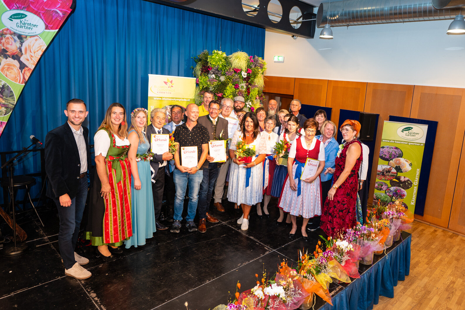 Blumenolympiade Kärnten: Preisträgerinnen und Preisträger mit Urkunden und Blumensträußen vor floraler Wand
