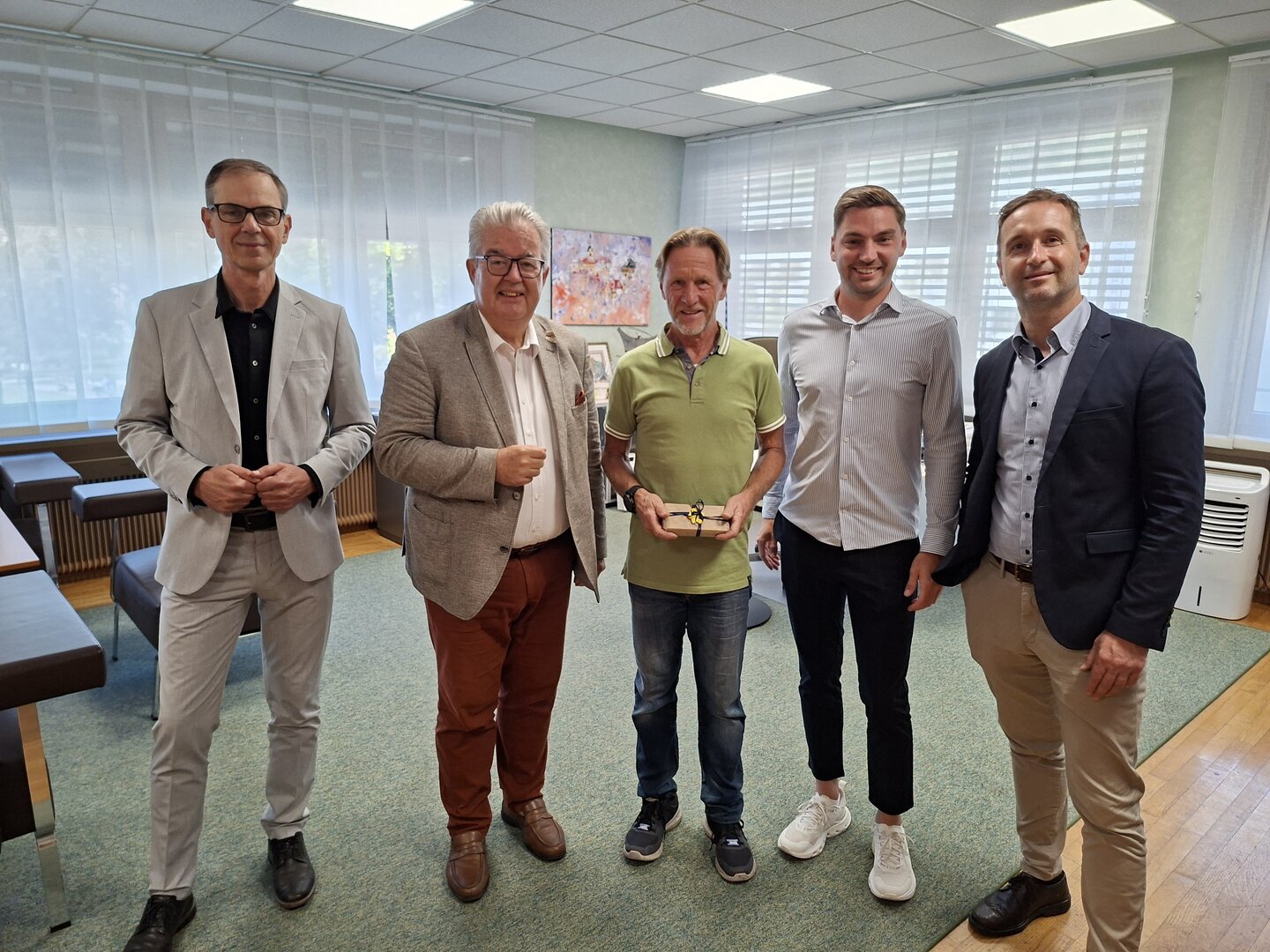 Fünf Männer in Anzügen stehen in einem Büro; einer hält ein Geschenk.